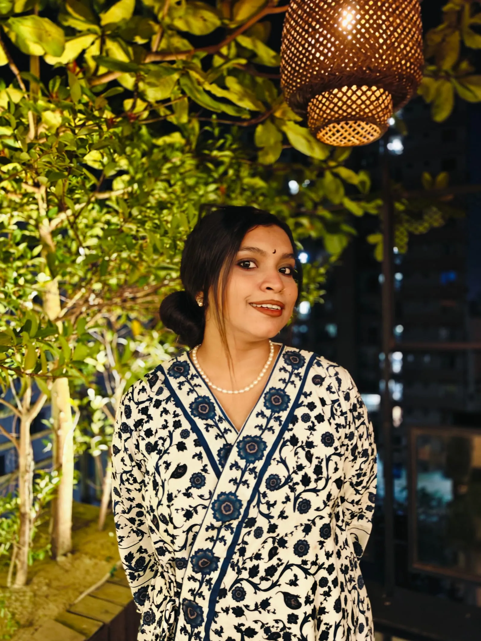 Shafrin Akter Chowdhury standing among potted trees on a rooftop terrace at night, wearing a blue block-printed kurta.