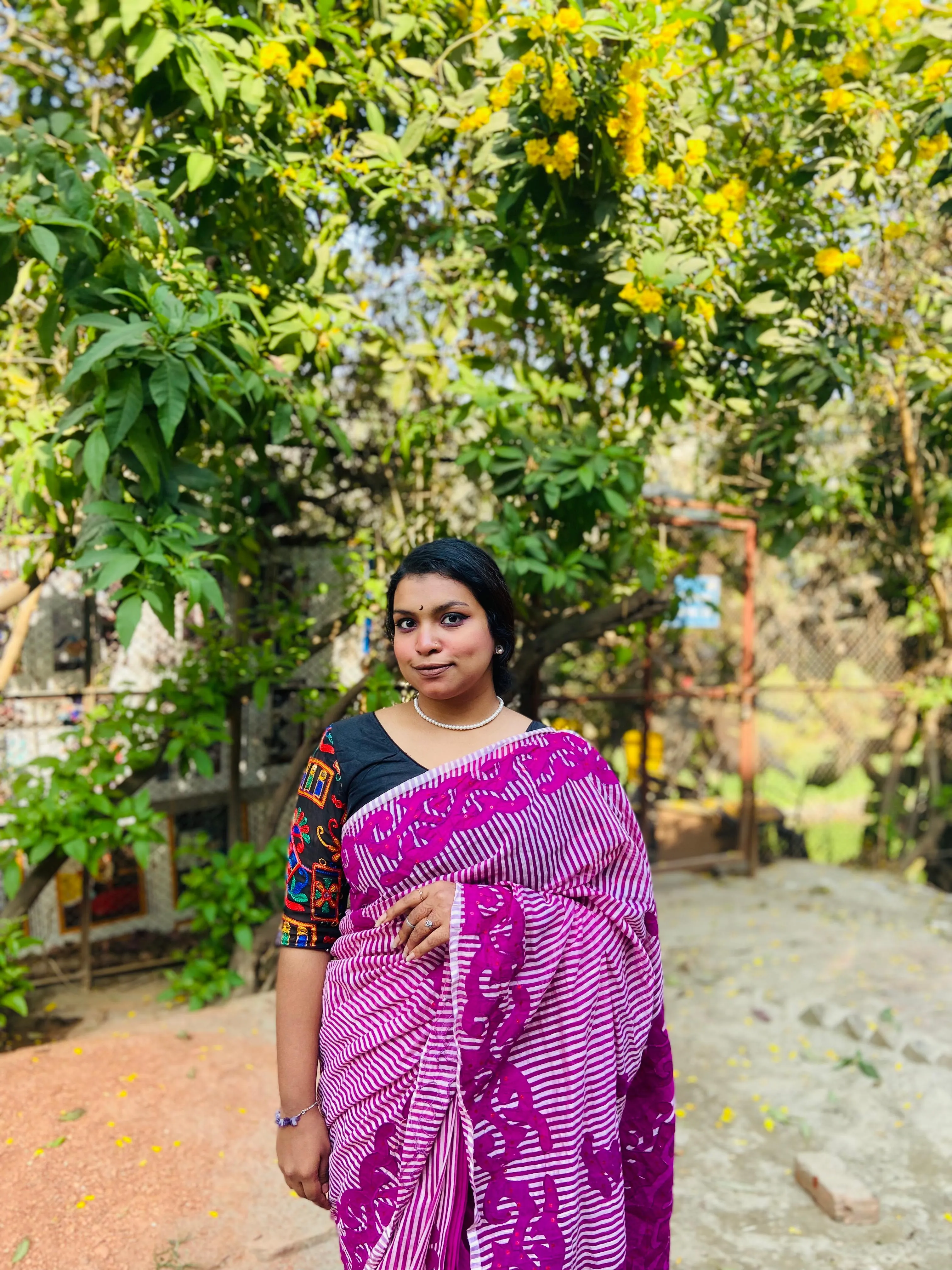 Shafrin Akter Chowdhury standing on a garden path beneath a flowering tree, wearing a magenta block-printed saree.
